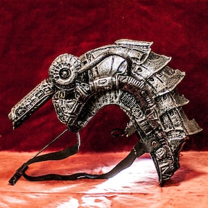 Pode incluir: Um capacete prateado, detalhado e texturizado com uma correia. O capacete tem um design futurista e parece ser feito de metal.