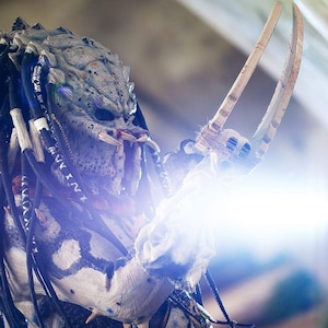 Pu&ograve; includere: Un costume da Predator con un designo a motivi bianco e nero. Il costume presenta una maschera con una mascella appuntita e una lunga treccia di dreadlocks bianchi e neri che fluiscono. Il Predator tiene un paio di lame lunghe e affilate.
