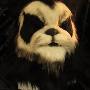 Può includere: Una maschera di pelliccia nera e bianca con un'espressione minacciosa. La maschera ha un naso e una bocca neri e pelliccia bianca intorno agli occhi.