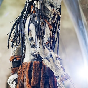 Pu&ograve; includere: Un costume da Predator con una maschera bianca e nera, dreadlock neri e una gonna di pelliccia marrone. Il costume tiene un'arma con una lama.