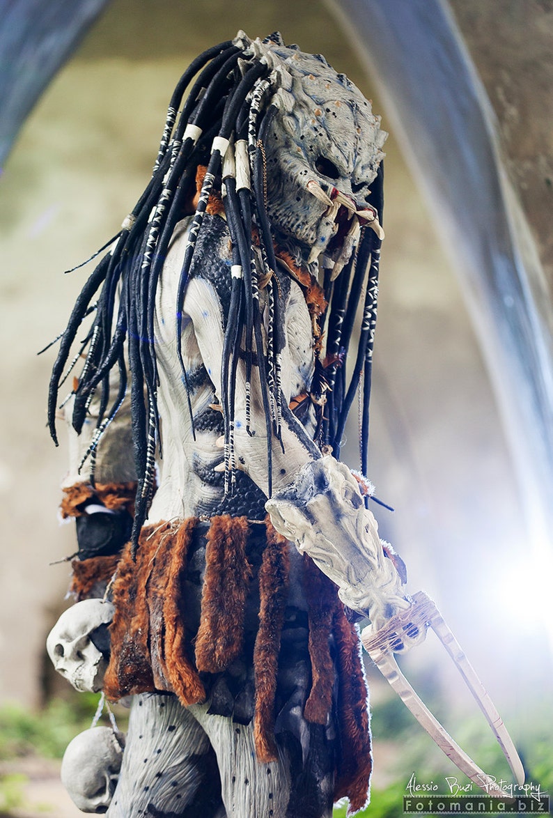 Pu&ograve; includere: Un costume da Predator con una maschera bianca e nera, dreadlock neri e una gonna di pelliccia marrone. Il costume tiene un'arma con una lama.