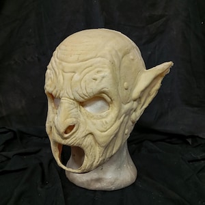 Op de afbeelding: Een beige goblinmasker met gedetailleerde gezichtskenmerken en puntige oren. Het masker is gemaakt van een zacht, buigzaam materiaal en heeft een gat voor de mond.
