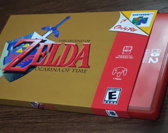 May include: A gold-colored video game box for The Legend of Zelda: Ocarina of Time featuring a sword and shield graphic, with a red Nintendo Switch game case partially inserted into a cardboard sleeve displaying N64 and ESRB logos.