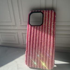 May include: A pink smartphone case featuring vertical rows of iridescent, light-reflecting crystals. The case has a black rectangular camera cutout at the top left corner and a textured surface covered in small, shimmering, bead-like embellishments.