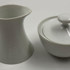 May include: A white porcelain creamer pitcher with a curved handle and a matching white sugar bowl featuring a round lid with a knob handle. Both pieces have a smooth, glossy finish and a simple, clean design for serving coffee or tea.