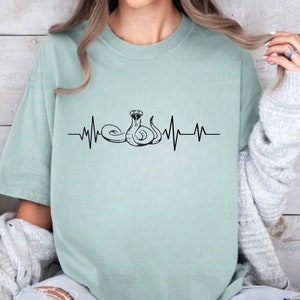 May include: A light green t-shirt with a black snake design and a heartbeat graphic. The snake is coiled and has its head raised. The heartbeat graphic is below the snake.