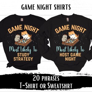 May include: Two black t-shirts with a game night theme. One shirt says "Game Night Most Likely To Study Strategy" and the other says "Game Night Most Likely To Host Game Night". Both shirts feature a cartoon image of two cats playing a board game.