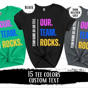 May include: Three t-shirts in different colors with the text "Our Team Rocks." The text is in pink, blue, and yellow on a white background. The t-shirts are in heather Irish green, black, and dark heather grey.