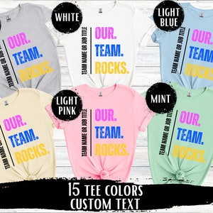May include: A graphic design for a t-shirt with 15 different color options. The design features the text "Our Team Rocks." in pink, blue, and yellow lettering.