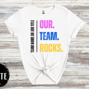May include: White t-shirt with the text "OUR. TEAM. ROCKS." printed in pink, blue, and yellow. The text is centered on the shirt and is surrounded by a black border.