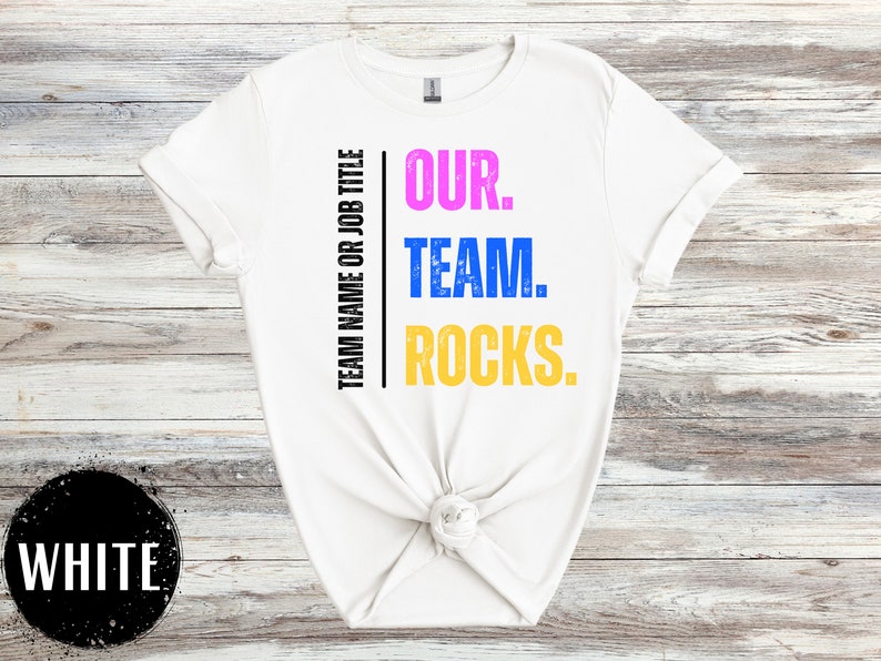 May include: White t-shirt with the text "OUR. TEAM. ROCKS." printed in pink, blue, and yellow. The text is centered on the shirt and is surrounded by a black border.