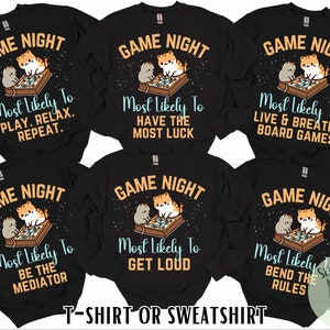 May include: Six black long-sleeve t-shirts with a graphic of two cats playing a board game. Each shirt has the text "Game Night" and a different phrase about board games. The phrases are "Most Likely To Play. Relax. Repeat.", "Most Likely To Have The Most Luck", "Most Likely To Live & Breathe Board Games", "Most Likely To Be The Mediator", "Most Likely To Get Loud", and "Most Likely To Bend The Rules".
