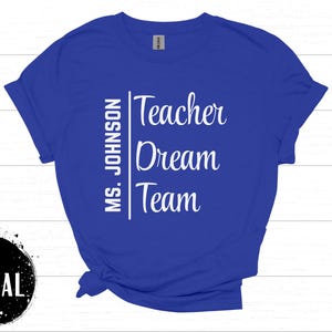 May include: Royal blue t-shirt with the words "Teacher Dream Team" in white script. The name "Ms. Johnson" is printed vertically on the left side. The shirt is displayed on a white wooden surface.