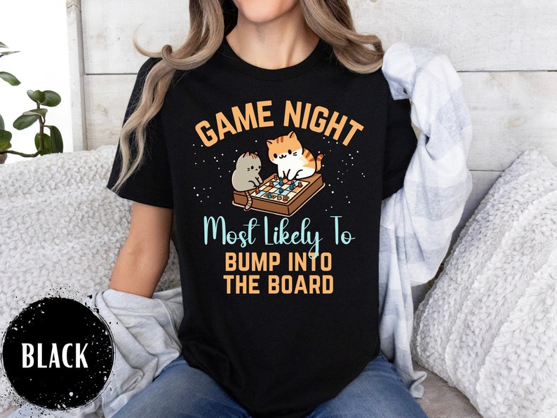 May include: Black t-shirt with a white graphic of two cats playing a board game. The text "Game Night Most Likely To Bump Into The Board" is printed on the shirt.