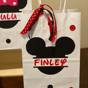 May include: White paper gift bag with black Mickey Mouse ears and the name "FINLEY" in red letters. The bag has red and white polka dot ribbon and black ribbon tied to the handles.