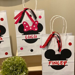 May include: Two white paper gift bags with black handles. The bags have red and black polka dots and feature a black and white silhouette of Mickey Mouse's head. One bag has a red bow and the name "Lulu" printed on it. The other bag has the name "Finley" printed on it.
