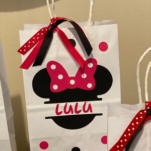 May include: White paper gift bag featuring a black and red Minnie Mouse design. The bag has red and white polka dot ribbon and black ribbon. The bag is personalised with the name "Lulu".