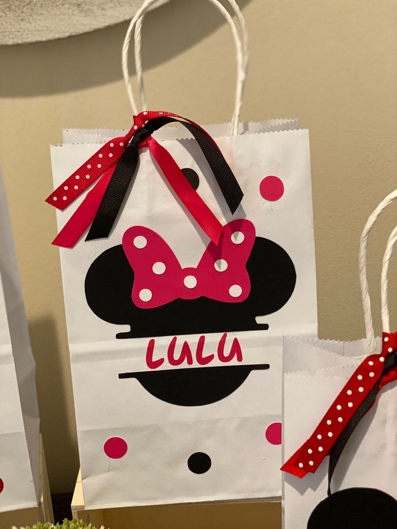 May include: White paper gift bag featuring a black and red Minnie Mouse design. The bag has red and white polka dot ribbon and black ribbon. The bag is personalised with the name "Lulu".