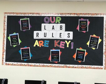 Op de afbeelding: Een zwart prikbord met een kleurrijk bord waarop staat "Our Lab Rules Are Key!" Het bord is gemaakt van afzonderlijke letters en is omgeven door kleinere borden met regels voor het lab. De borden hebben een zwarte achtergrond met witte tekst en kleurrijke randen.