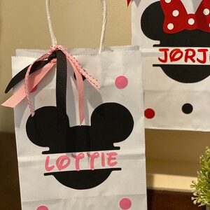 May include: White paper gift bag with black Mickey Mouse ears and the name "Lottie" printed on it. The bag has pink and black polka dots and a pink and black ribbon.