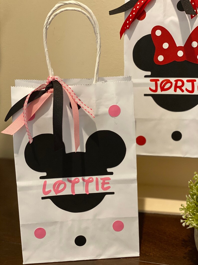 May include: White paper gift bag with black Mickey Mouse ears and the name "Lottie" printed on it. The bag has pink and black polka dots and a pink and black ribbon.