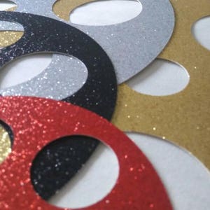 May include: A set of five round, glittery paper circles with cut-out centers. The circles are in red, gold, black, and silver.