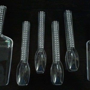 May include: A collection of clear plastic serving utensils, including scoops and spoons. The handles are decorated with a grid pattern of small, clear embellishments. The items are arranged against a dark background.