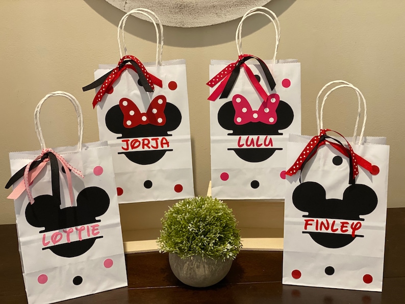 May include: Four white paper gift bags with black handles and red and black polka dots. Each bag features a black and white silhouette of Mickey Mouse's head with a red bow and the name "LULU", "JORGJA", "FINLEY", and "LOTTIE" printed on the front.