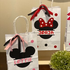 May include: Two white paper gift bags with black handles and red and black polka dots. The bags feature black Mickey Mouse ears with a red bow on top. The bags are personalised with the names "Lottie" and "Jorja" in pink lettering.