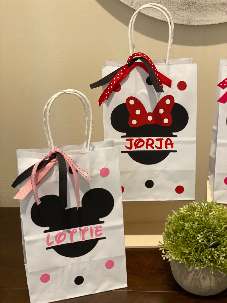 May include: Two white paper gift bags with black handles and red and black polka dots. The bags feature black Mickey Mouse ears with a red bow on top. The bags are personalised with the names "Lottie" and "Jorja" in pink lettering.
