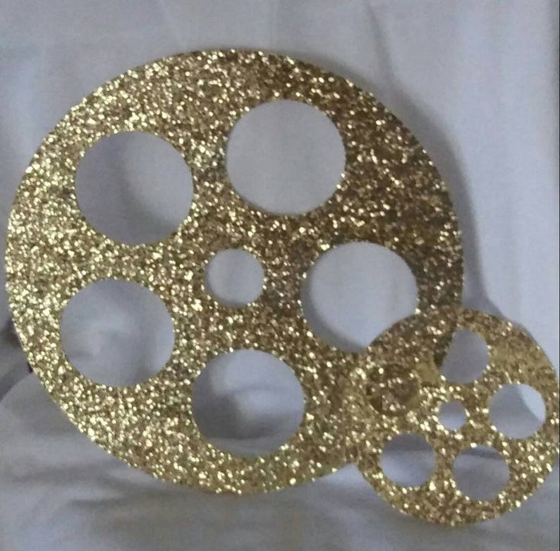 May include: Two gold glitter film reel ornaments with cut-out circles. The larger reel has five circles and the smaller reel has three circles.
