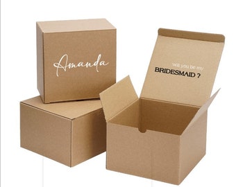 May include: Three brown cardboard boxes. The top box has the name "Amanda" written in white script. The open box has the text "Will you be my BRIDESMAID?" printed on the inside.