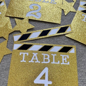 Puede incluir: Tarjeta de número de mesa de purpurina dorada con rayas negras y blancas, que se asemeja a una claqueta de cine. La tarjeta dice "TABLE 4" en letras blancas.