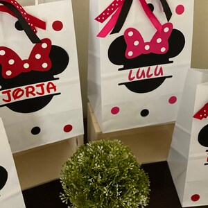 May include: Three white paper gift bags with black Mickey Mouse ears and red bows. The bags are decorated with red and white polka dots and have names written on them: "Jorja", "Lulu", and "Finley".