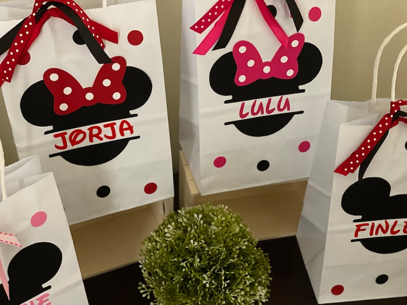 May include: Three white paper gift bags with black Mickey Mouse ears and red bows. The bags are decorated with red and white polka dots and have names written on them: "Jorja", "Lulu", and "Finley".