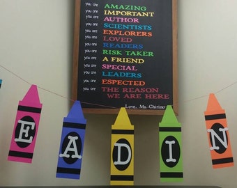 Puede incluir: Una pancarta colorida deletrea "READING" con cada letra en una hoja de papel con forma de crayón separada. La pancarta está colgada en una pared con una pizarra detrás. La pizarra tiene una lista de afirmaciones positivas sobre los estudiantes, incluyendo "Eres increíble", "Eres importante", "Eres un autor", "Eres un científico", "Eres un explorador", "Eres amado", "Eres un lector", "Eres un tomador de riesgos", "Eres un amigo", "Eres especial", "Eres un líder", "Eres esperado", "Eres la razón por la que estamos aquí". La pizarra está firmada "Love, Ms. Chirino".