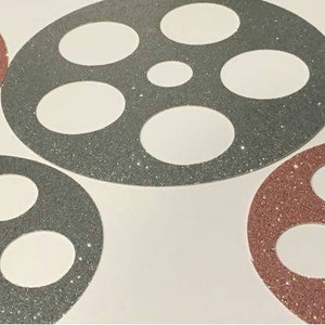 May include: Four decorative film reel shapes, two in a dark gray glitter and two in a rose gold glitter. The film reels are on a white background.