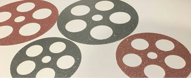 May include: Four decorative film reel shapes, two in a dark gray glitter and two in a rose gold glitter. The film reels are on a white background.