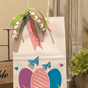 May include: White paper gift bag with a green, brown, and blue ribbon and a pink tag that says "Happy Easter". The bag is decorated with three paper Easter eggs, two butterflies, and green grass.