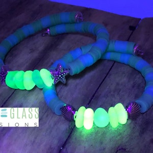 May include: Two green and blue striped bracelets with glow-in-the-dark sea glass beads and silver starfish charms. The bracelets are on a wooden surface. "SEA GLASS VISIONS" is written on the surface.