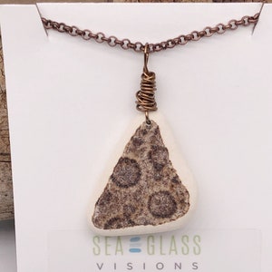 May include: A copper chain necklace with a white triangular pendant featuring a brown and white speckled pattern. The pendant is attached to the chain with copper wire. The necklace is displayed on a white card with the text "Sea Glass Visions by Cathy Kelly".