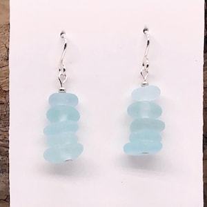 May include: A pair of dangle earrings featuring light blue sea glass pieces stacked on silver hooks. The earrings are displayed on a white card with the text "SEA GLASS VISIONS by Cathy Kelly" in green and blue. The background includes driftwood.