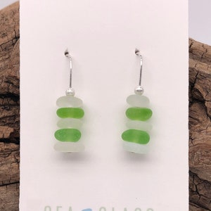 May include: A pair of silver dangle earrings with three stacked pieces of sea glass in shades of green and white. The earrings are on a white card with the text "Sea Glass Visions by Cathy Kelly".