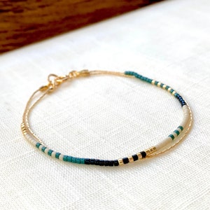 Puede incluir: Un delicado brazalete dorado con un diseño en capas de cuentas blancas, negras y azul verdoso.