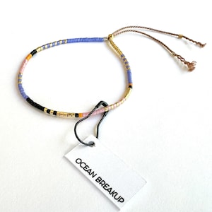 May include: A purple, pink, black, and gold beaded bracelet with a brown cord and a tag that reads "Ocean Breakup".