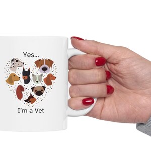 May include: White ceramic mug with a heart-shaped design of various dog breeds. The mug has the text "Yes... I'm a Vet" printed on it. The mug is held by a person with red nail polish.