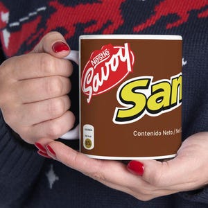 May include: A white ceramic mug with a brown background featuring the Nestle Savoy logo in red and white, and the word "San" in yellow. The mug is held by a person with red nail polish.