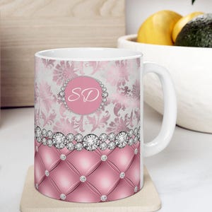 May include: A white ceramic mug with a pink and silver design. The mug features a damask pattern with a pink circle in the center that says "SD". The bottom of the mug has a pink quilted design with silver rhinestones.