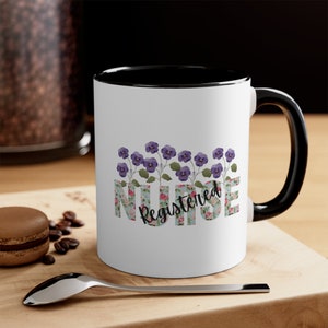 May include: White ceramic mug with a black interior and handle. The mug features the text "Registered Nurse" in a floral design with purple flowers. The mug is sitting on a wooden surface.