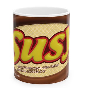 May include: A white ceramic mug with a brown and yellow design. The mug features the word "Susj" in yellow bubble letters with a red outline. The mug also has text in Spanish: "GALLETA RELLENA CON CREMA SABOR A CHOCOLATE".